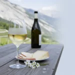 A glass of pale Savoy white wine on a rustic table with alpine vineyards and mountains in the background.