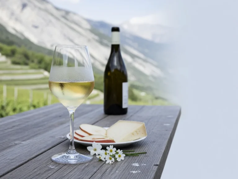 A glass of pale Savoy white wine on a rustic table with alpine vineyards and mountains in the background.
