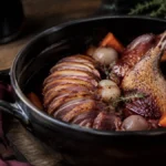 Tender duck served with pearl onions and vegetables in a glossy red-wine sauce beside a rustic cocotte.