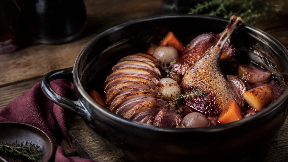 Tender duck served with pearl onions and vegetables in a glossy red-wine sauce beside a rustic cocotte.