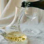 Crystal decanter being filled with pale golden white wine poured from a dark green wine bottle, captured in elegant editorial